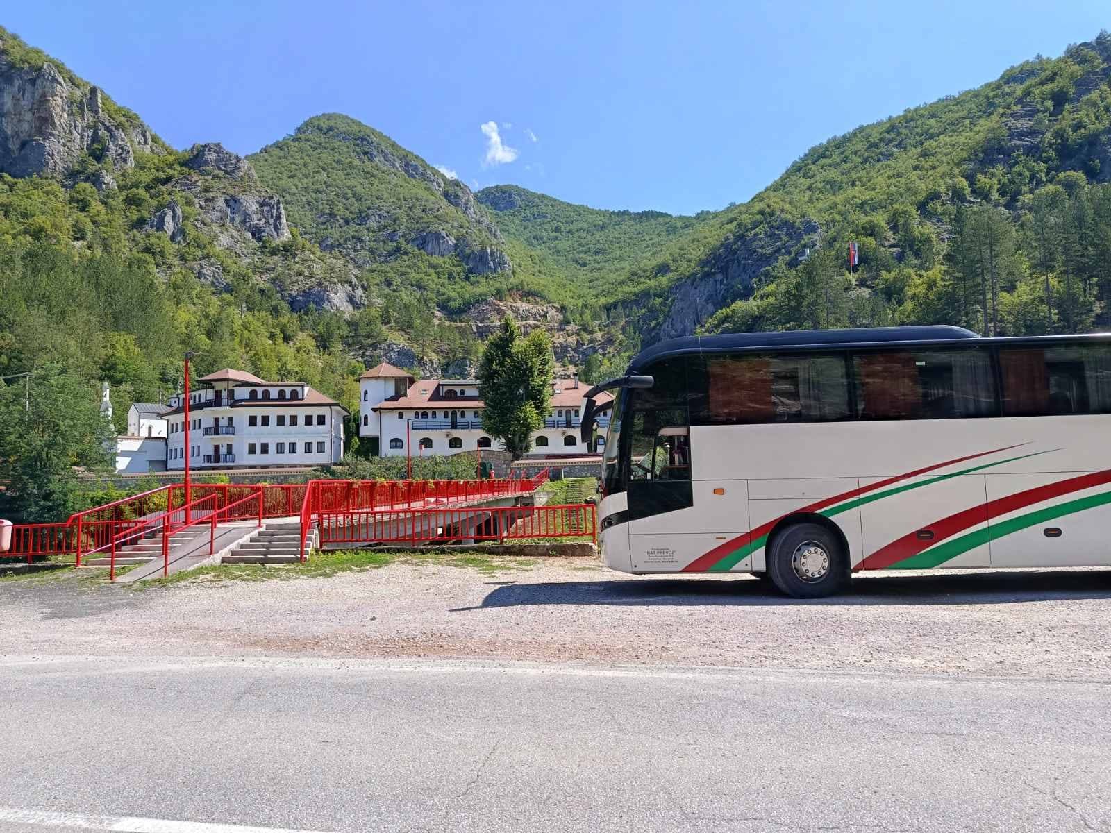 Beli autobus ispred manastira u klisuri sa crvenim mostom